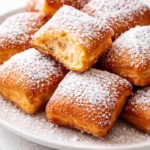 Golden brown, airy New Orleans beignets piled on a plate, generously dusted with powdered sugar and emitting a light vanilla aroma.