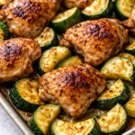 Golden and juicy baked chicken pieces alongside tender zucchini slices, lightly charred at the edges, on a baking sheet.