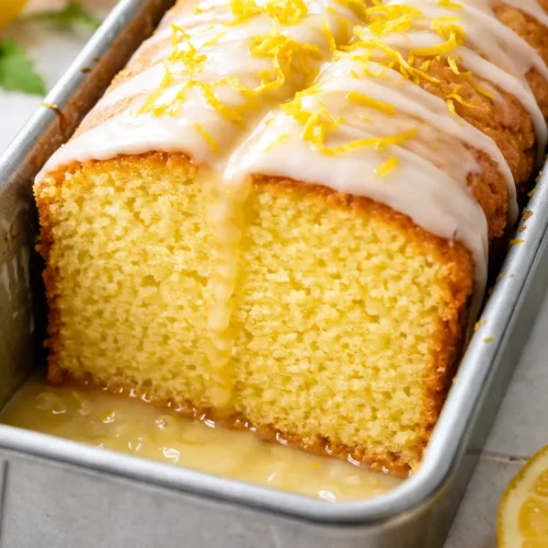 A slice of soft, moist lemon cake with glossy lemon glaze and bright yellow zest on a white plate, with a fork alongside.
