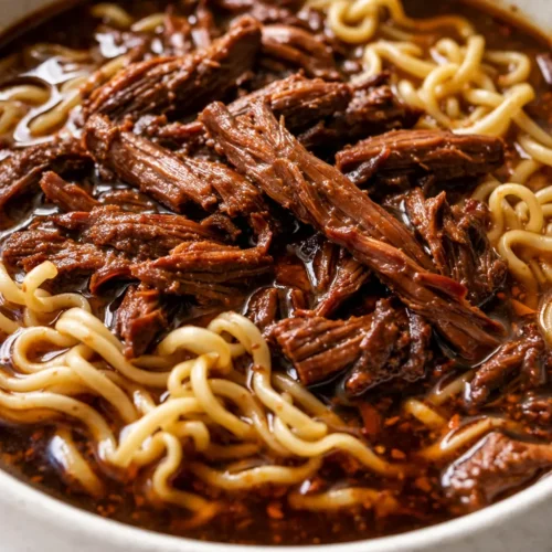 Bowl of deep, rich slow cooker beef ramen with savory broth, tender beef slices, soft-boiled egg, green onions, and sesame seeds.