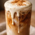 Tall glass of creamy iced apple crisp nondairy cream chai latte, with frothy oat milk top and ice cubes, amber caramel color