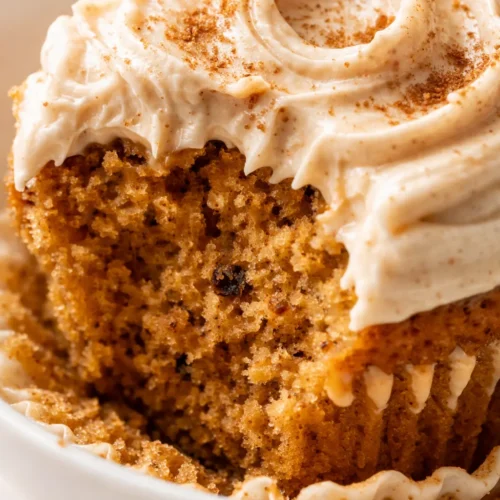 Swoops of brown sugar cream cheese frosting atop moist pumpkin cupcakes on a rustic wooden stand, dusted with cinnamon