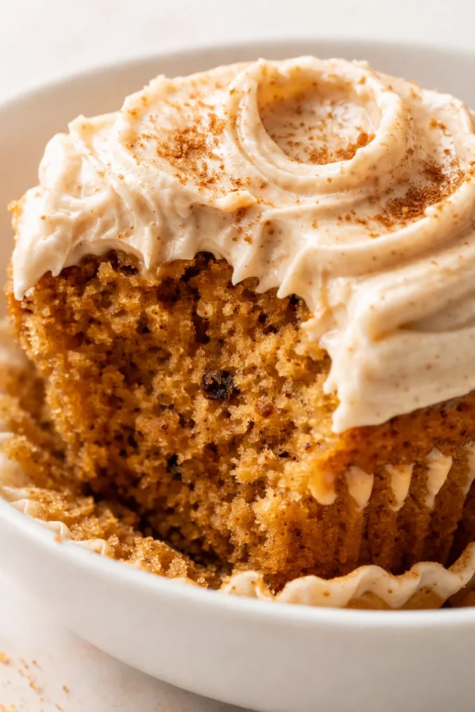 Swoops of brown sugar cream cheese frosting atop moist pumpkin cupcakes on a rustic wooden stand, dusted with cinnamon