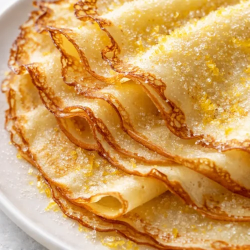 Lemon sugar crepes stacked on a plate, showing tender texture, crisp golden edges, and a light dusting of sugar.